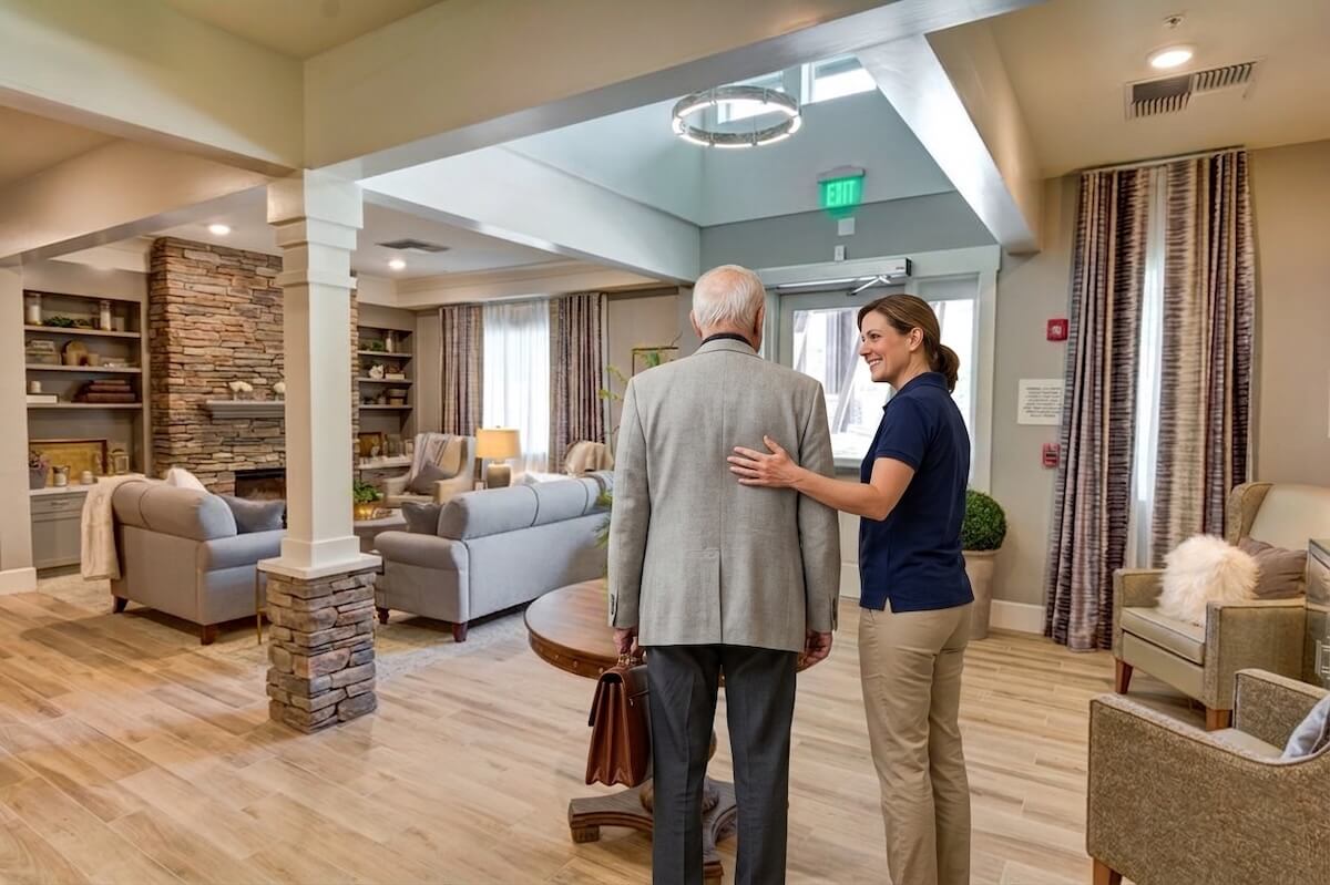 Caregiver guiding elderly man through welcoming senior living lounge
