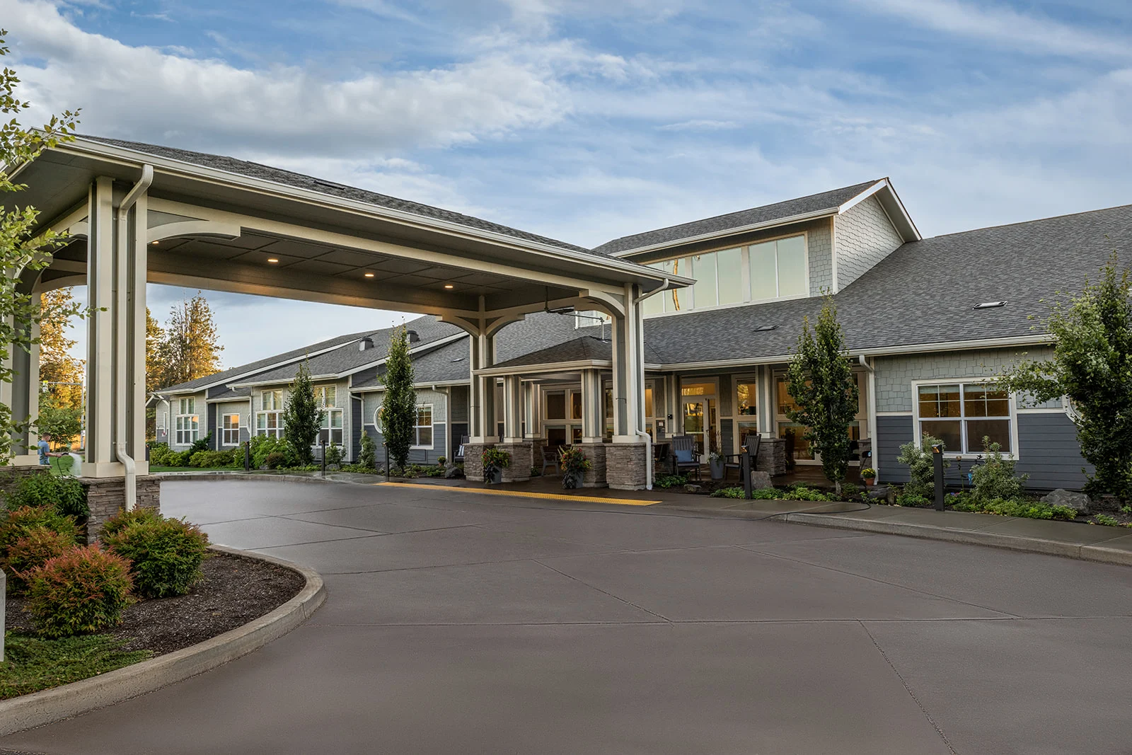 Exterior of a modern senior living community building with a covered driveway entrance, surrounded by trees, shrubs, and landscaping under a partly cloudy sky.