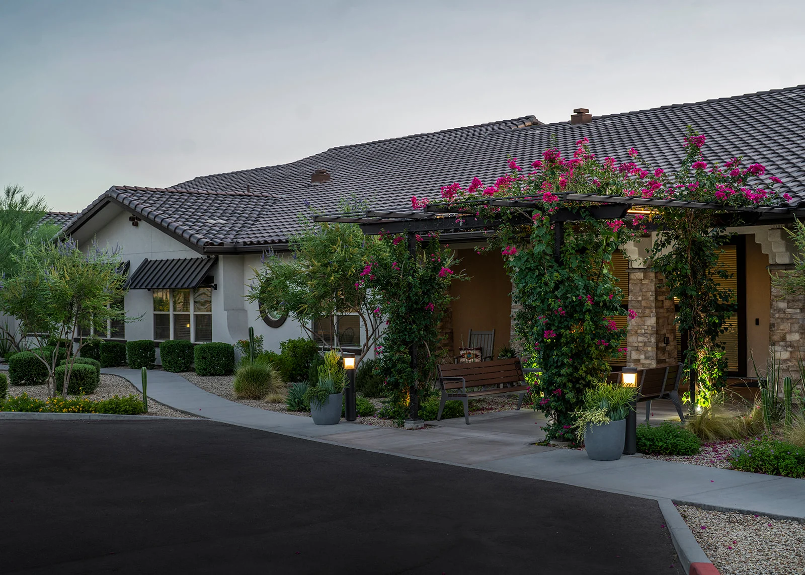 Suburban house with tiled roof, landscaped garden, flowering vines covering shaded seating area near entrance, benches, and lamp posts along sidewalk.